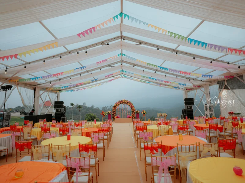 Brightly decorated event tent interior with yellow and orange tables, gold chairs, colorful bunting, and a floral arch over a central walkway.